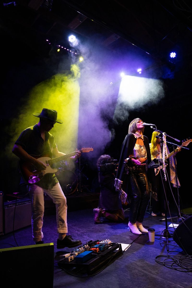 A band performing on stage under dramatic lights, with a videographer filming them.