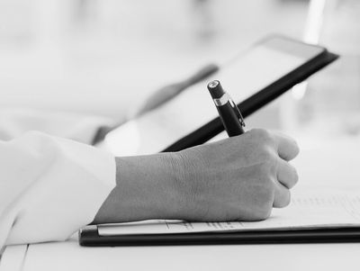 Close-up of a hand writing with a pen on paper, holding a tablet in the background.