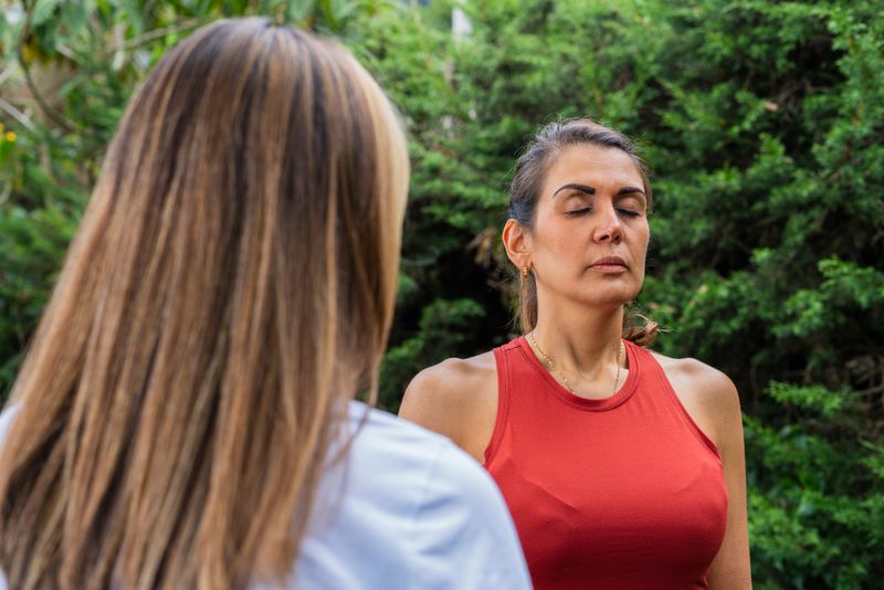 Average age 35-year-old Latina woman with blonde hair dressed comfortably stands in the garden of her open-air meditation office with her patient 30-year-old Latina woman who is dressed comfortably and attends meditation and yoga retreats where she is taught to control your breathing