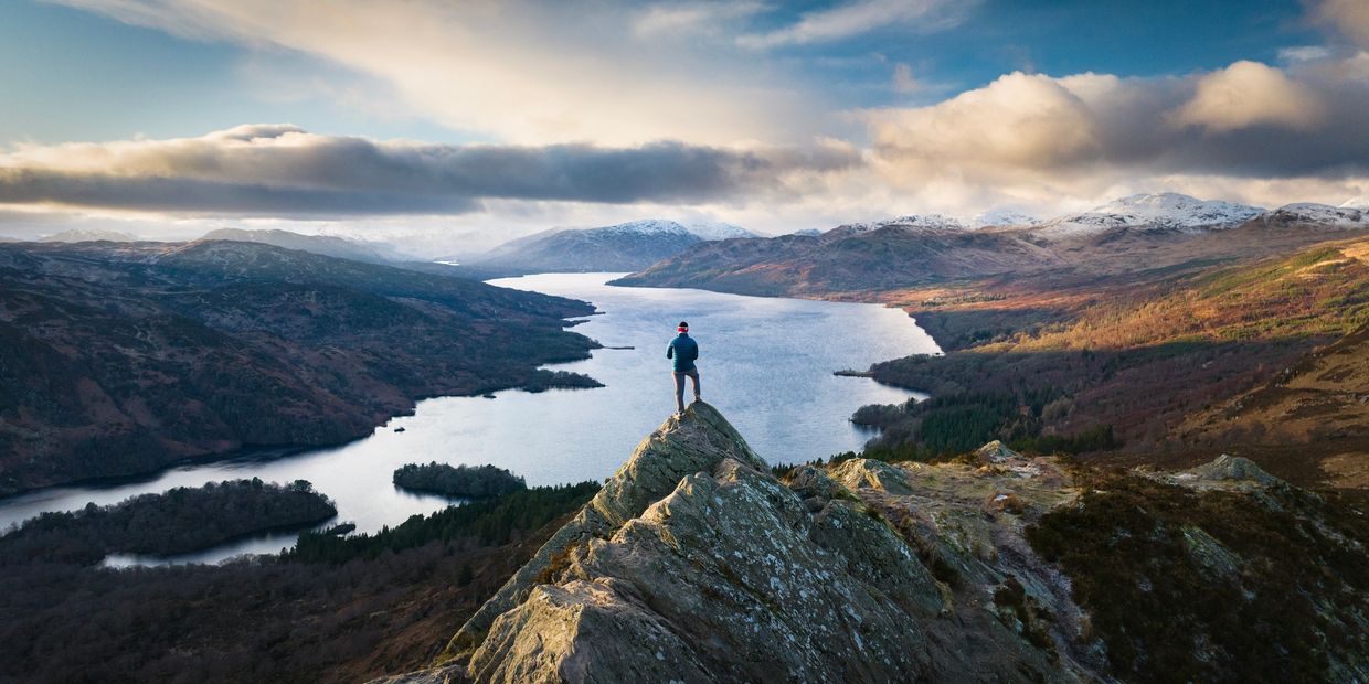 A person stands atop a rocky peak overlooking a serene lake and mountain range.