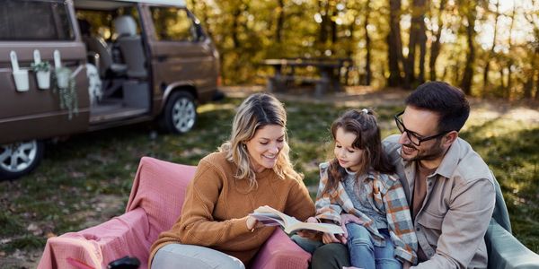 A family enjoying a book outdoors near a camper van in a forest setting.