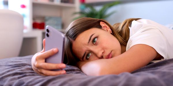 Young woman lying on bed, looking at her phone with a sad expression.