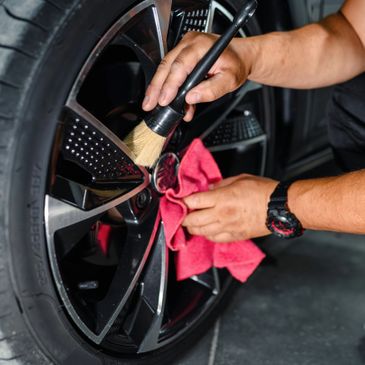 Person cleaning a car's black alloy wheel with a brush and red cloth.