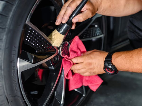 Person cleaning a car's black alloy wheel with a brush and red cloth.