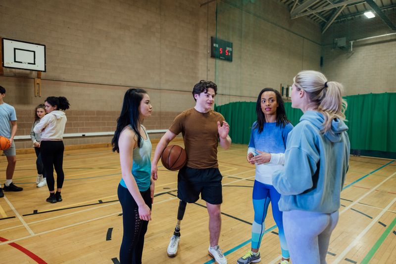 A wide-angle shot of a group of teenagers playing basketball on an indoor sports court. They are standing in two teams and planning their game approach. One of the male players is an amputee and has a bionic leg.