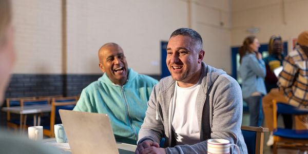 Two men laughing and chatting in a casual indoor setting with a laptop.