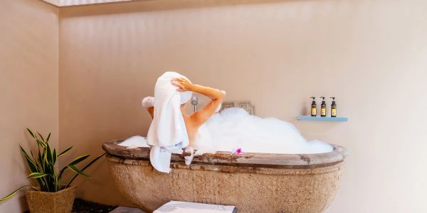 Person relaxing in a bubble bath with a towel on their head in a cozy bathroom.