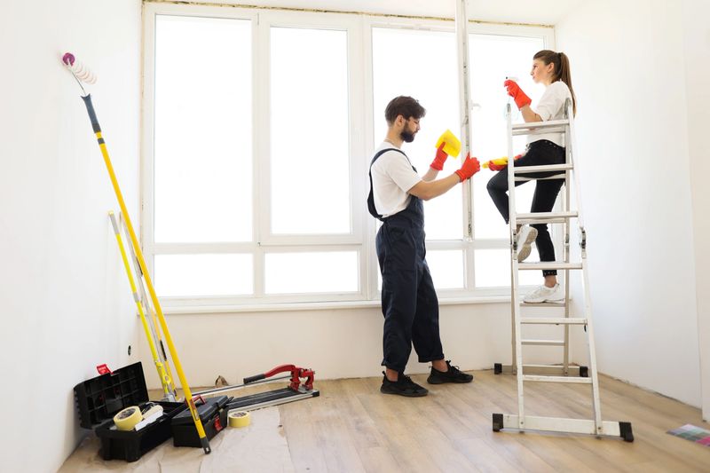 Young family of two, pretty woman sitting on ladder and bearded man household chores, they wash the window in their house along with cleaning agent and yellow house cleaning rags.