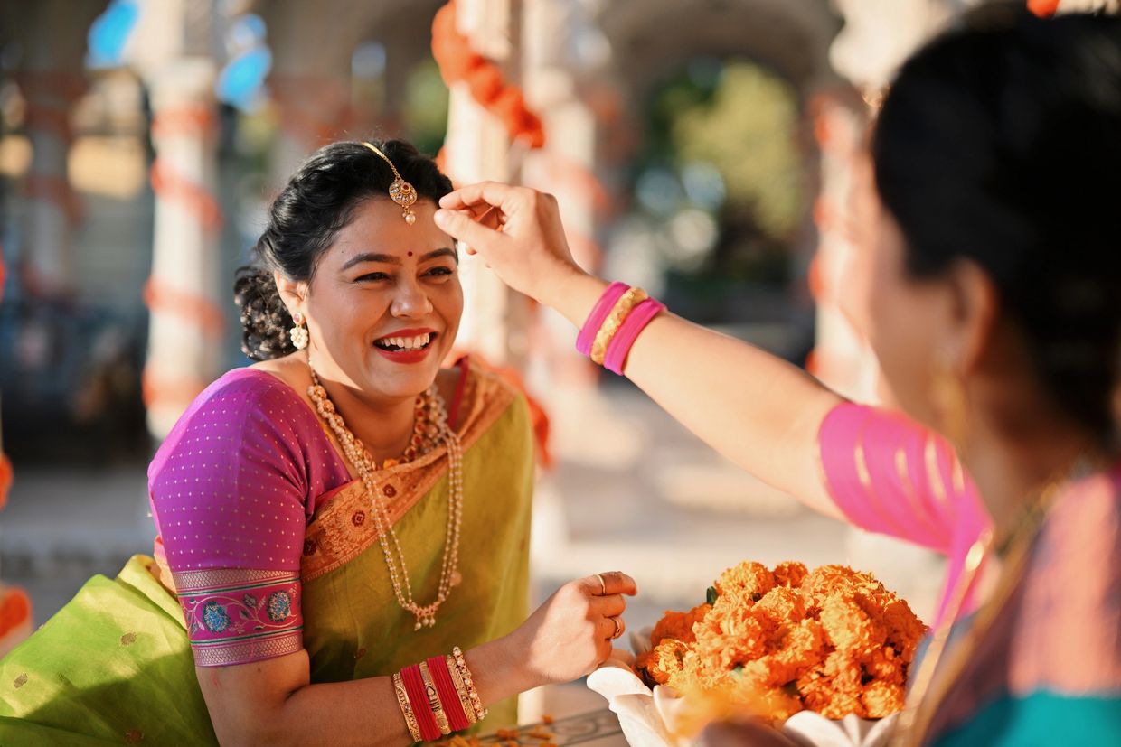 Two South Asian women in colorful traditional attire and jewelry, smiling at a festive gathering.