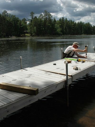 Man repairing a wooden dock by the lake under a cloudy sky.