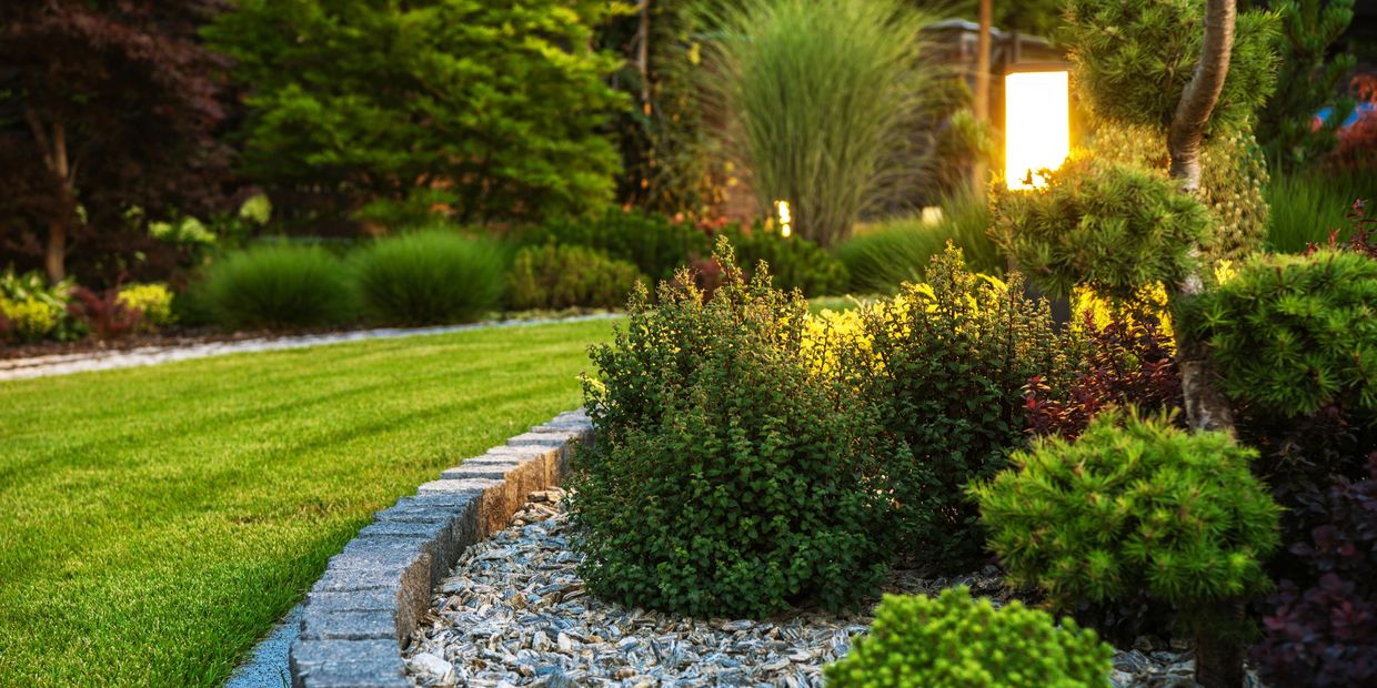 Well-maintained garden with trimmed bushes, stone border, and glowing lamp.
