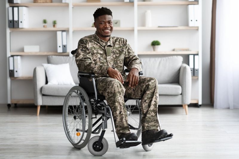 Happy young black in camouflage uniform soldier sitting in wheelchair and smiling at camera, having rehabilitation at clinic for injured veterans, copy space, full length shot