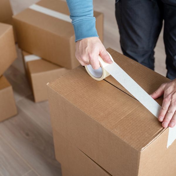 Person sealing a cardboard box with tape among several packed boxes.
