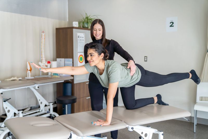 A young female client kneels on an assessment table as she stretches out and maintains her balance.  Her female therapist is spotting and supporting her as she walks her through the exercise.