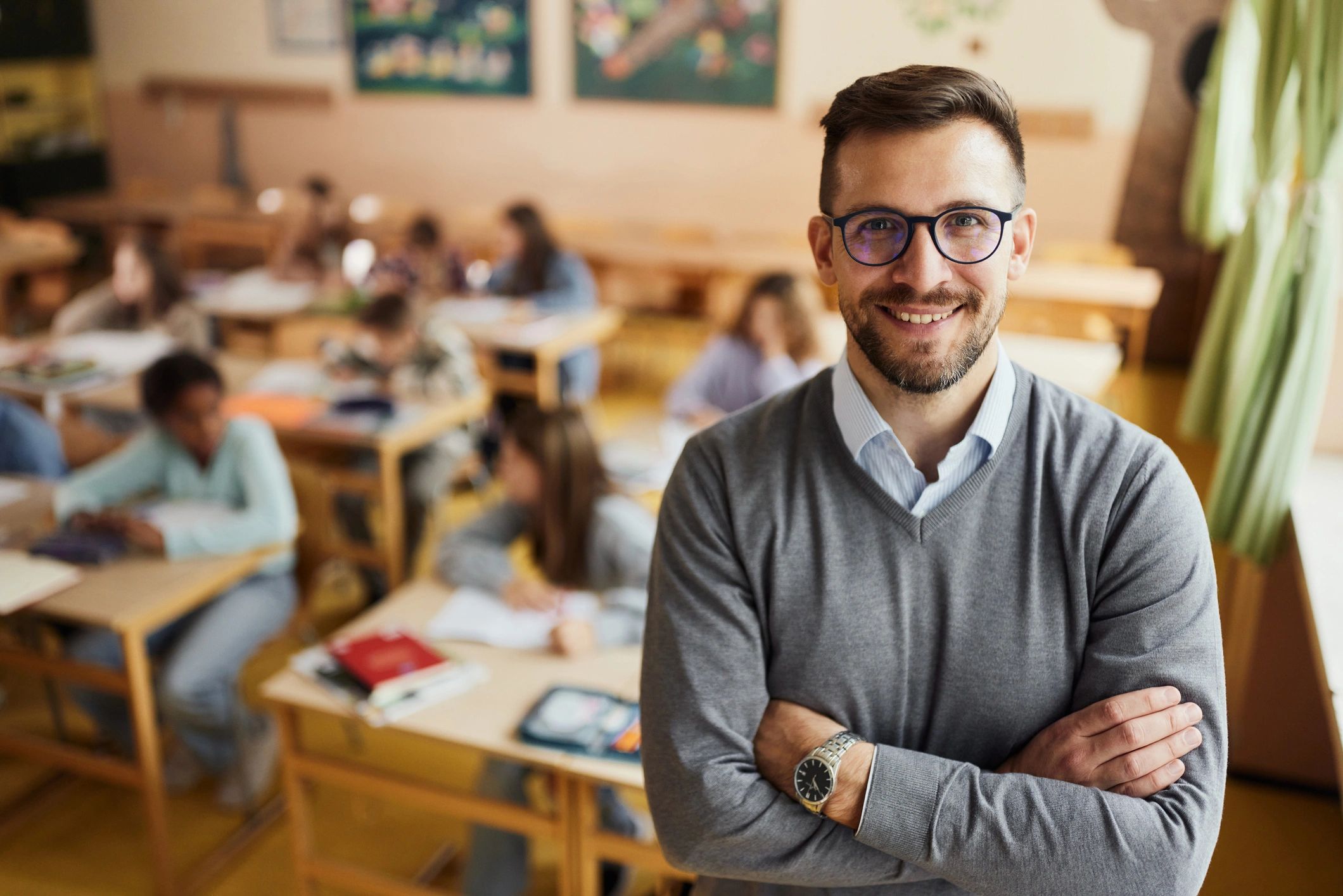A teacher wearing glasses stands in front of his students, who are sitting at their desks.