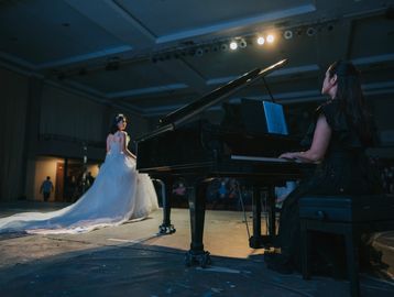A pianist accompanies a singer in a flowing white gown on stage during a performance.