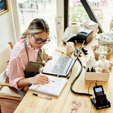 Woman in apron working on laptop and writing notes at a wooden table.