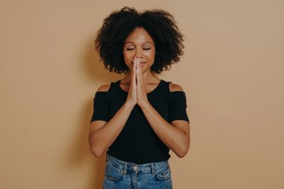 Woman with eyes closed and hands in prayer gesture, standing against a beige background.