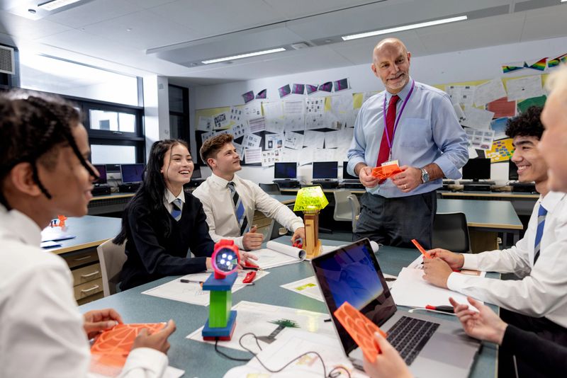 A low angle medium close-up of a group of students who are friends sitting in the classroom and working on a science technology project. They are working in smaller groups to brainstorm for their project.
