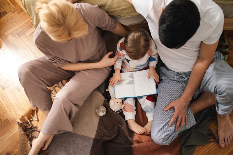 Directly above photo of a little girl reading a fairy tale while sitting with her mother and father on the bedroom floor.