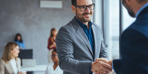 Hombres de negocios estrechándose la mano con colegas al fondo.