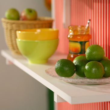 Fresh limes on a plate with colorful bowls and a basket of apples in the background.