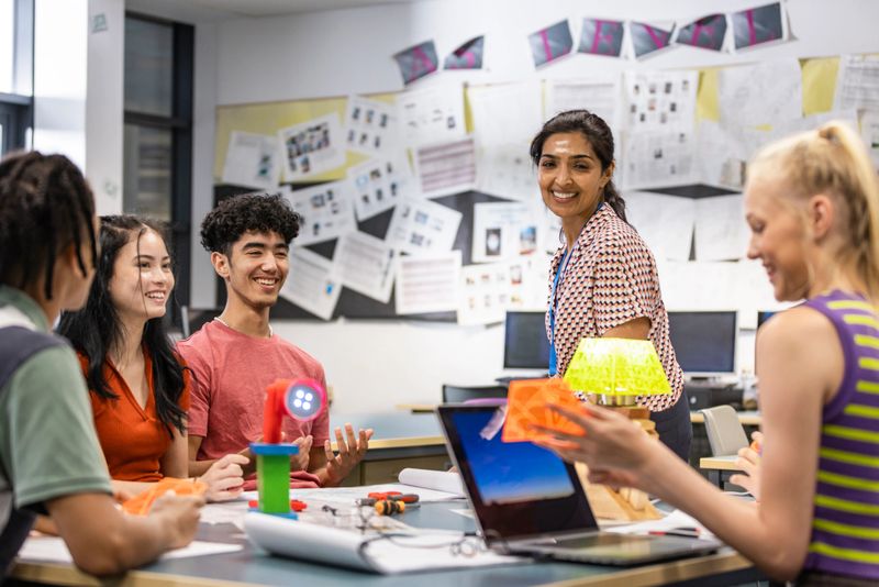 A shot of a group of secondary school students with their teacher in class, discussing their work. They are all wearing casual clothing and smiling, sitting in a classroom in a school in Gateshead, England.