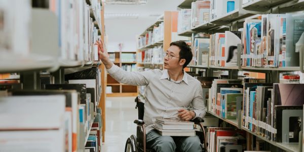 A man in a wheelchair reaching for a book in a library aisle.