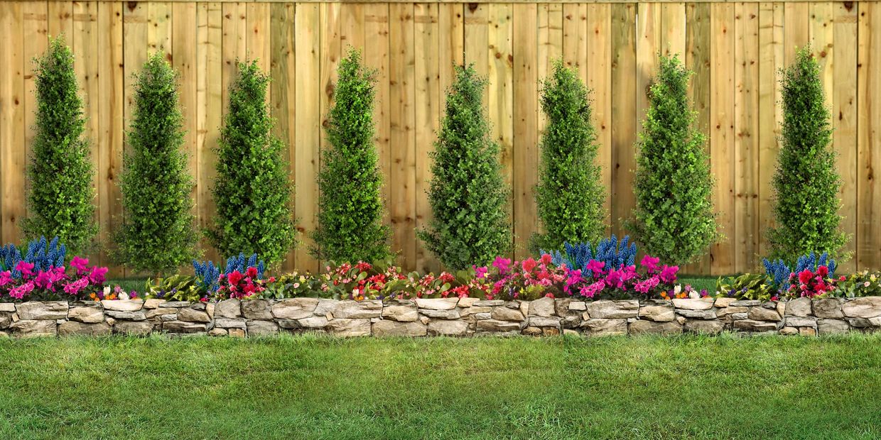 A neatly landscaped garden with tall green shrubs and colorful flowers in front of a wooden fence.