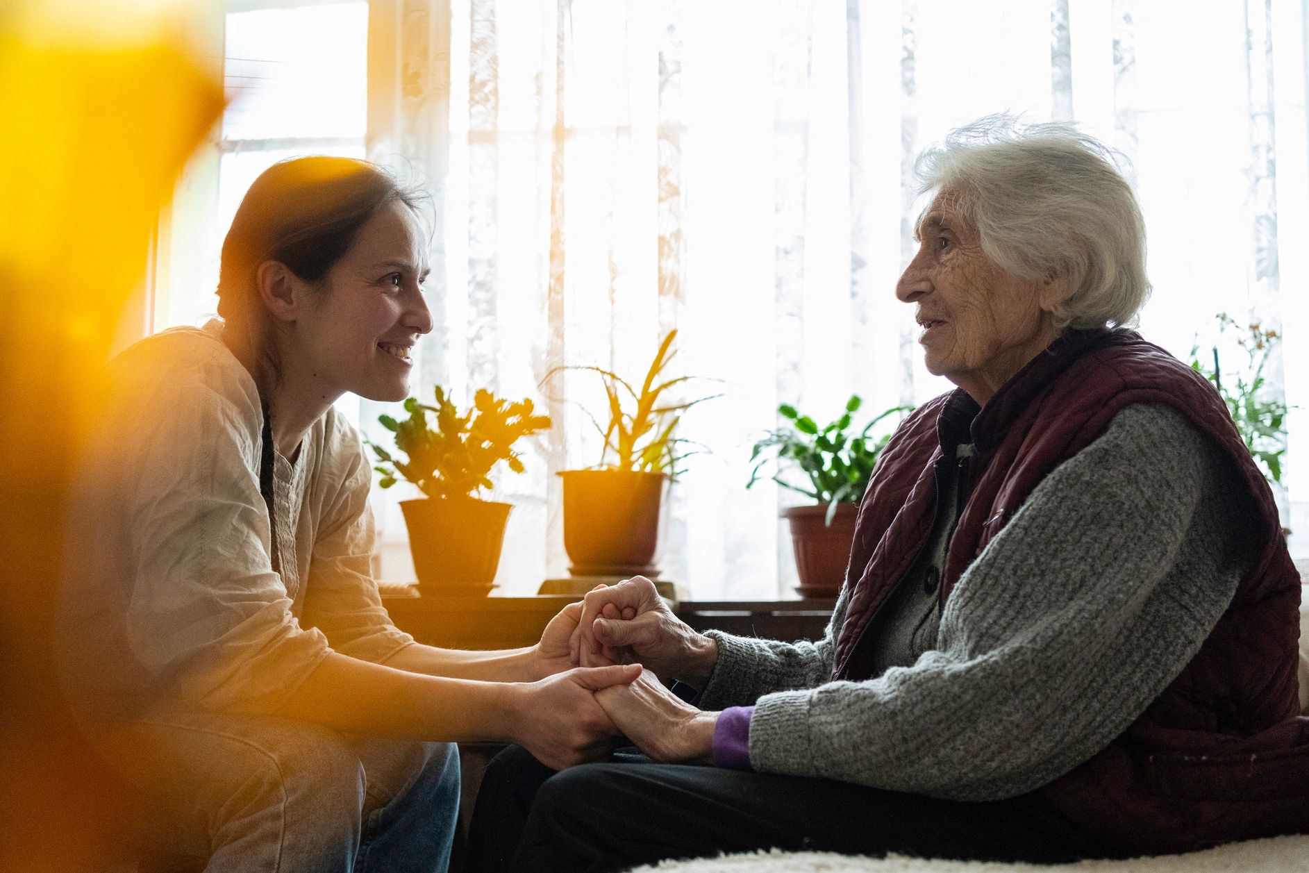 Young woman holding hands and smiling at an elderly woman in a cozy room with plants.