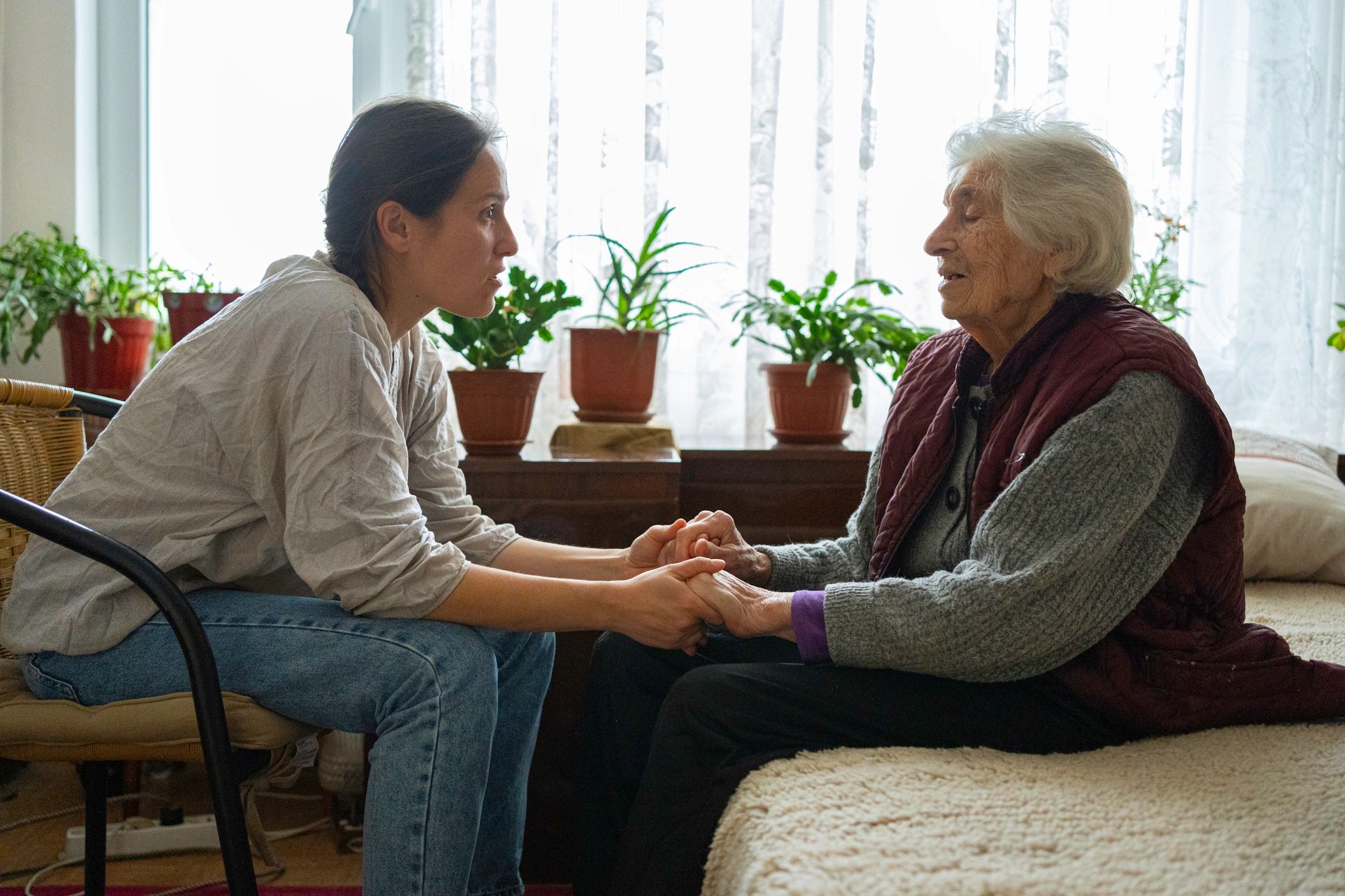 Two women holding hands and talking in a cozy room with plants.