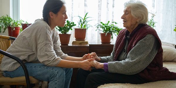 A young woman holds hands and talks with an elderly woman in a cozy room.