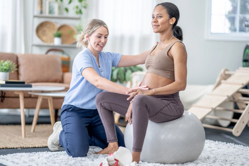 A female physiotherapist works with a pregnant woman during an appointment.  The woman is dressed comfortably and sitting on a yoga ball as they work through various exercises.