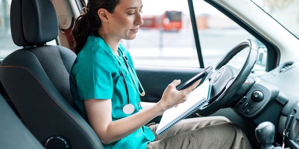 A healthcare professional in scrubs sitting in a car, using a phone and notebook.