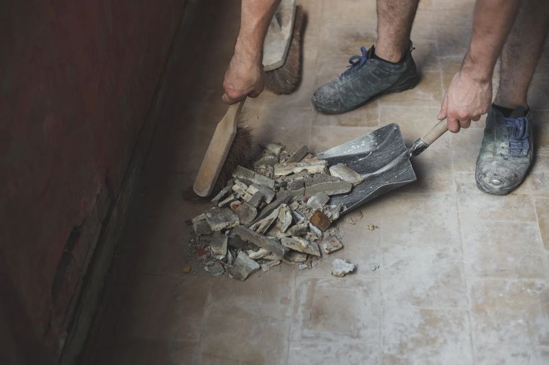Young caucasian unrecognizable man sweeping construction debris with a small whisk on an old dustpan from a dirty tiled floor, close-up side view. The concept of cleaning and installing windows, construction work, house renovation.