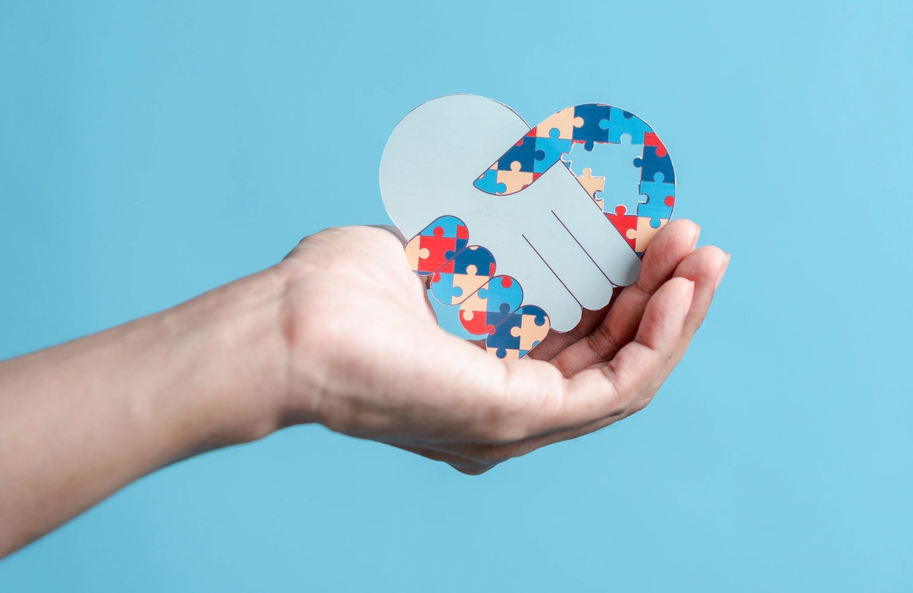 Hand holding a puzzle piece shaped like a handshake with autism colors.