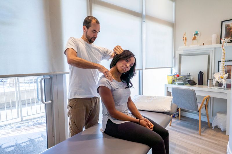 A young woman of African decent, sits up on the edge of a table as her Chiropractor carefully gives her an adjustment.   She is dressed casually and has her head tilted to the side as her Chiropractor walks her through the adjustment.  The Chiropractor is standing behind the patient as he moves the patients head and shoulder gently into position.
