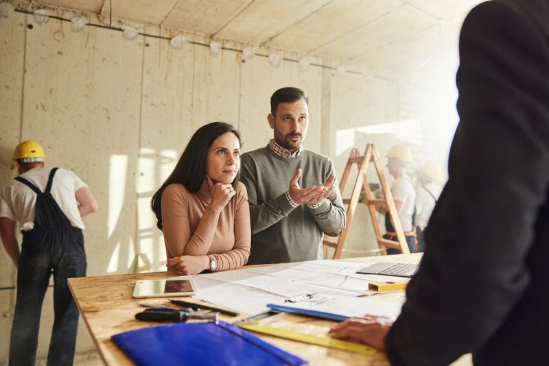 Young couple communicating with an architect while examining blueprints at construction site. Manual workers are in the background.