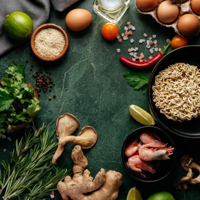 Overhead view of fresh ingredients including eggs, shrimp, noodles, and spices on a dark surface.