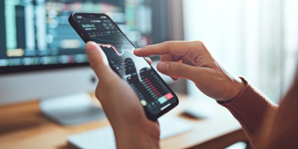 Person analyzing stock market data on a smartphone with a computer screen in the background.