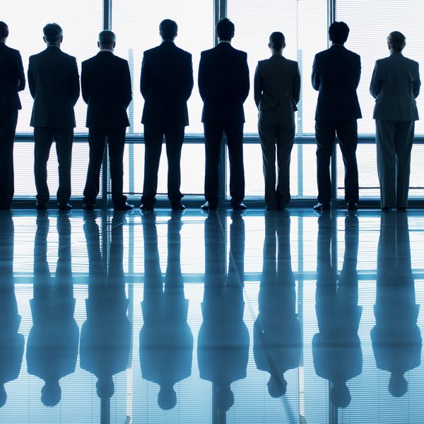 Eight business professionals standing in front of a large window, their reflections visible on the shiny floor.