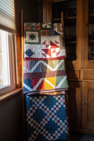 Colorful quilts hanging on a ladder near a window and wooden cabinet.