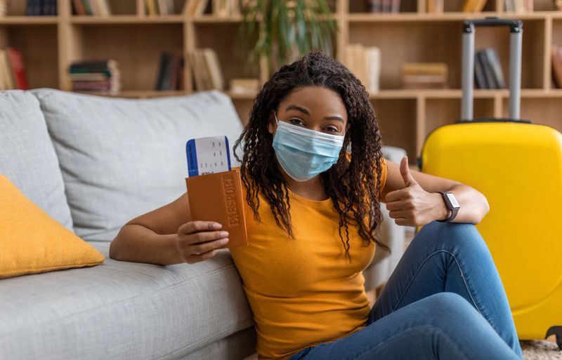 Choose safe tours. Happy black lady in protective mask with passport and tickets showing thumb up, ready to go on summer vacation trip. Traveling and tourism concept