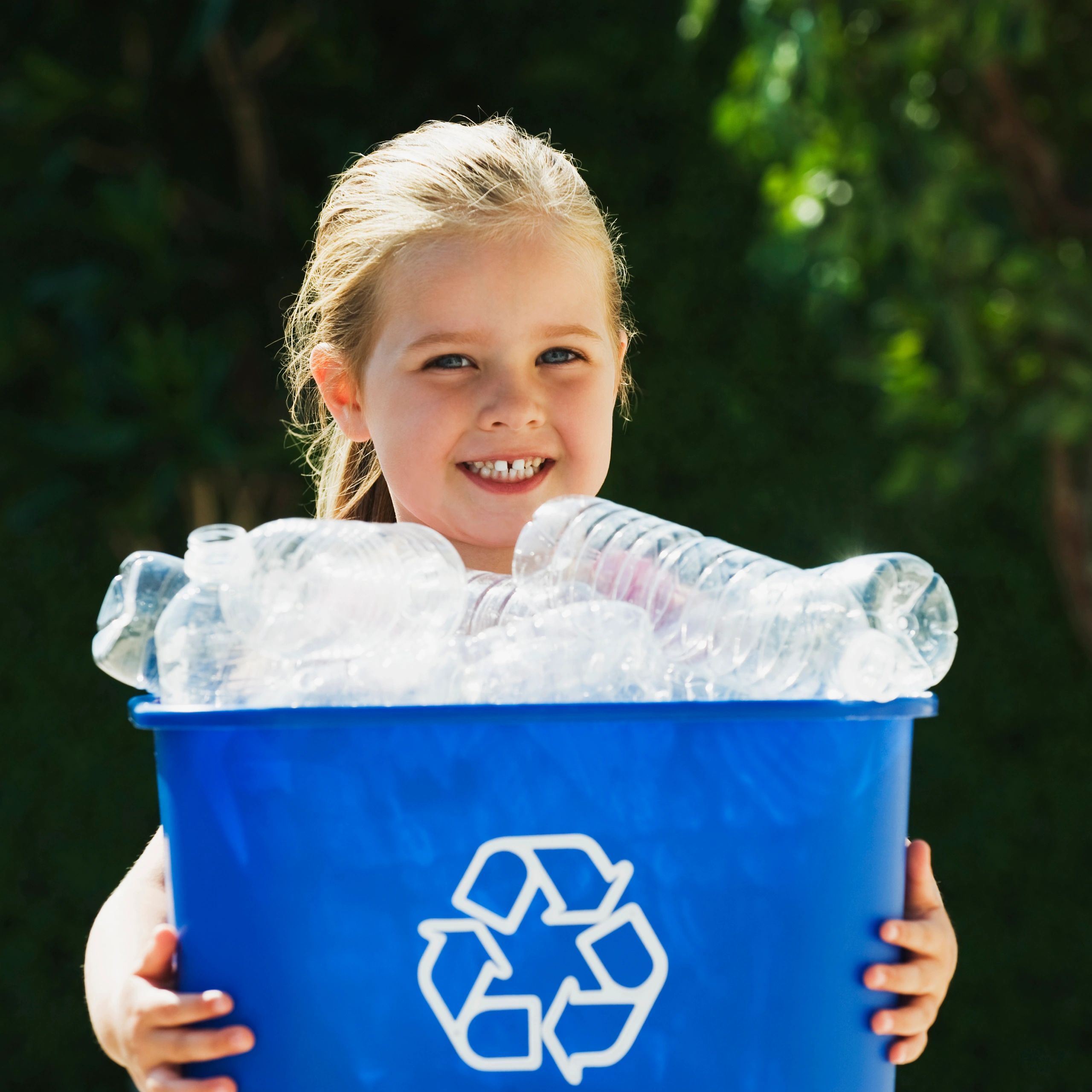 Smiling girl holding a blue recycling bin full of plastic bottles outdoors.