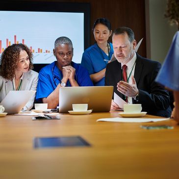 Medical professionals and a businessman discuss data around a laptop in a meeting room.