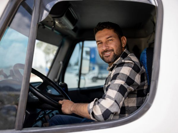 Smiling man behind the wheel of a truck wearing a checkered shirt.