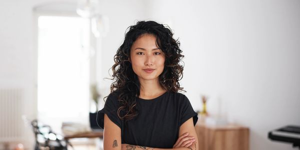 Confident young woman with curly hair and tattoos, arms crossed, standing indoors.