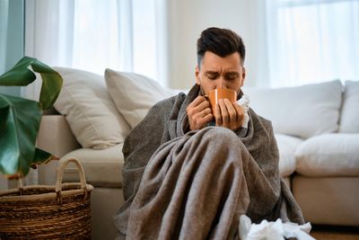 An ill man who is drinking tea from an orange cup, wrapped in a grey blanket and holding tissues.