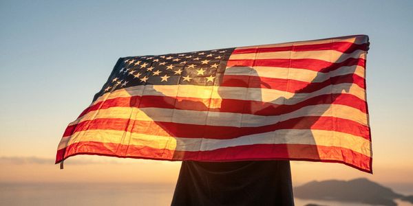 Person holding a translucent American flag at sunset with mountains in the background.