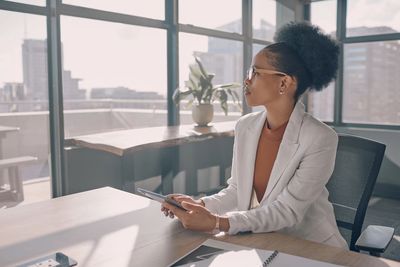 A woman in a white blazer holding a tablet, looking out an office window.